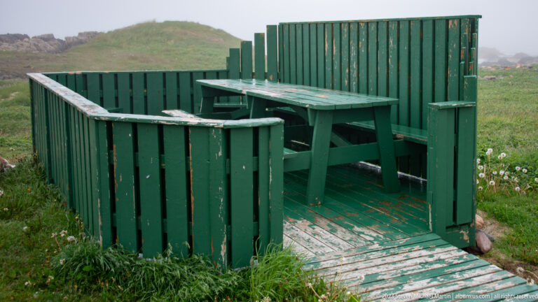 Green wooden wind shelter enclosing bench seating and a small table beside a boardwalk.