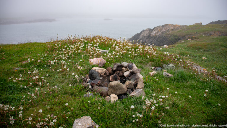 Small circular ring of rounded stones in a green coastal meadow filled with white seedheads.