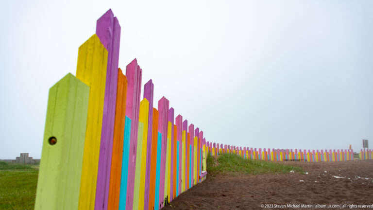 Low-angle view along bright multicolored wooden fence posts receding beside a foggy dark beach.
