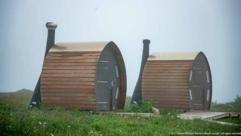 Two curved wooden pod shelters with black end walls, narrow windows, and stovepipes in dense fog.