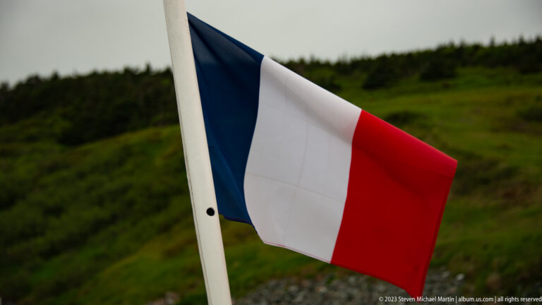 French blue-white-red tricolour flying from a pole against a green coastal hillside.