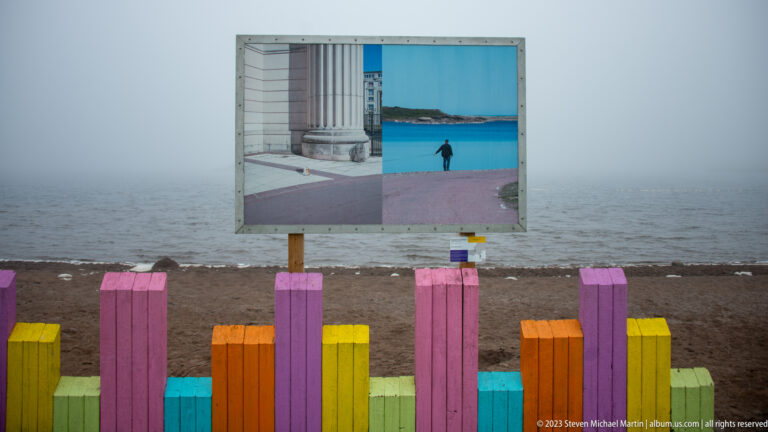 Outdoor panel with two photographic images standing behind a colorful timber fence on a foggy beach.
