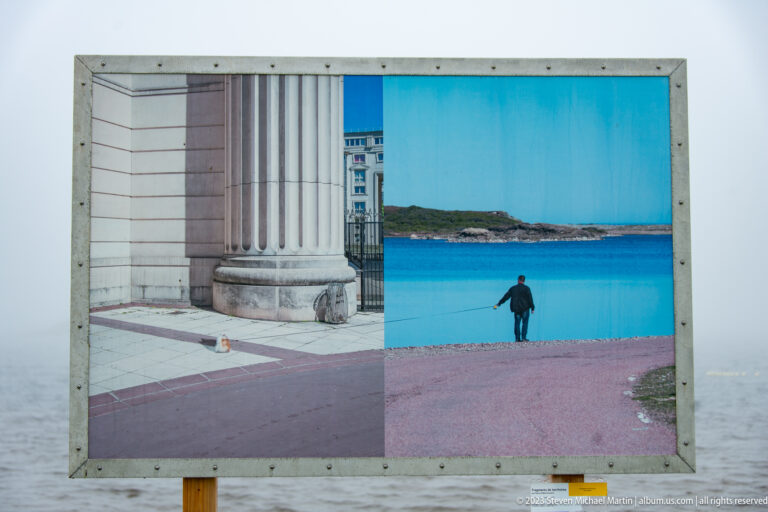 Outdoor framed panel displaying classical columns on the left and a person beside blue coastal water on the right.