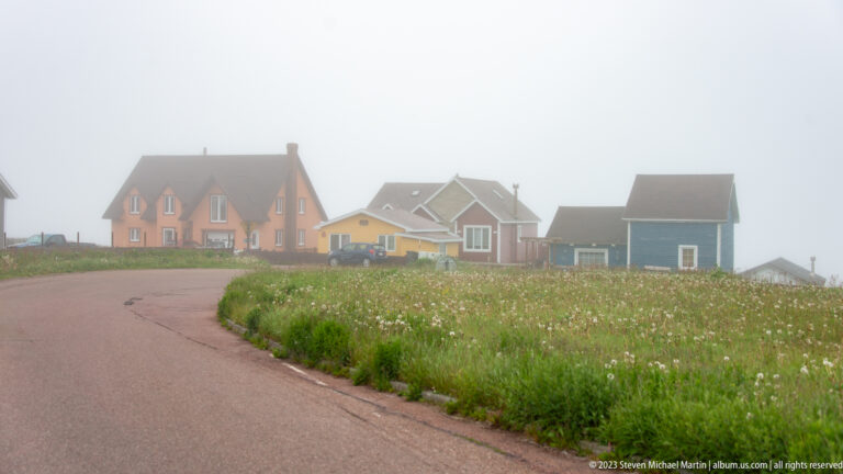 Curving paved road beside colorful detached houses and tall meadow grass in dense fog.