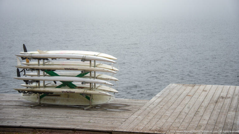 Several long white paddle craft stacked horizontally on a metal rack beside a wooden deck and gray water.
