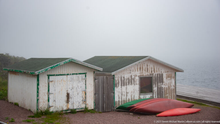 Two weathered white wooden sheds beside the water with red and green paddle craft lying in front.