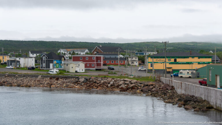 Small waterfront settlement with colorful houses, commercial buildings, rocky shore, and misty green hills.