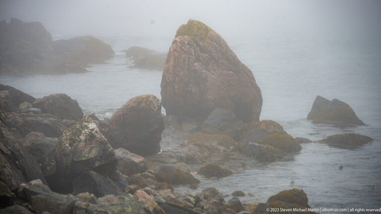Large pointed sea stack rising among smaller coastal boulders in gray water and dense fog.
