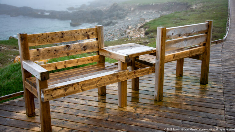 Two weathered wooden benches facing a small table on a wet deck beside a coastal boardwalk.