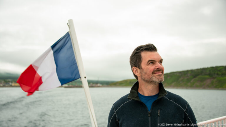 Man standing on a ferry deck beside a French flag, with wake and green shoreline behind him.
