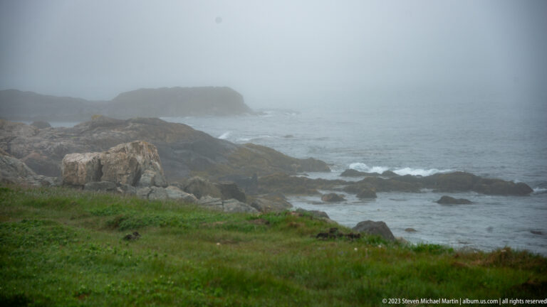 Rocky shoreline with low grass, breaking waves, and a distant headland fading into dense fog.