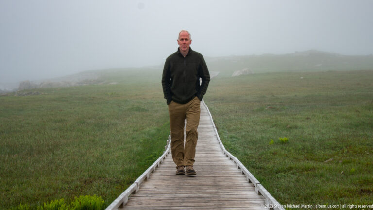 Man walking toward the camera on a straight wooden boardwalk through fog-covered green coastal terrain.