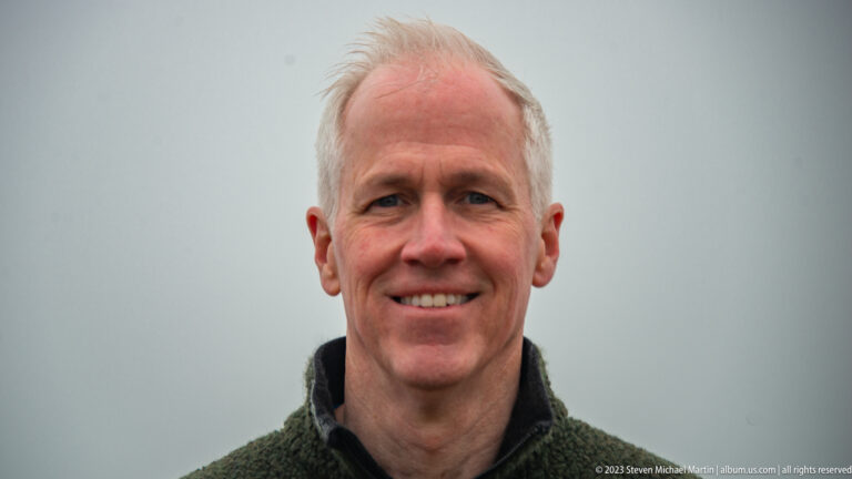 Close portrait of a smiling gray-haired man wearing a dark fleece against a pale foggy background.
