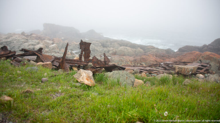 Rusting iron wreckage scattered among rocks and grass on a foggy shoreline, with the sea faintly visible beyond.