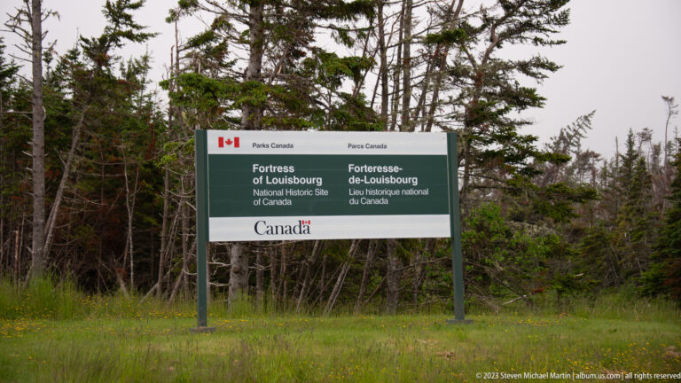 Bilingual Parks Canada entrance sign for Fortress of Louisbourg NHS standing beside a wooded roadside.
