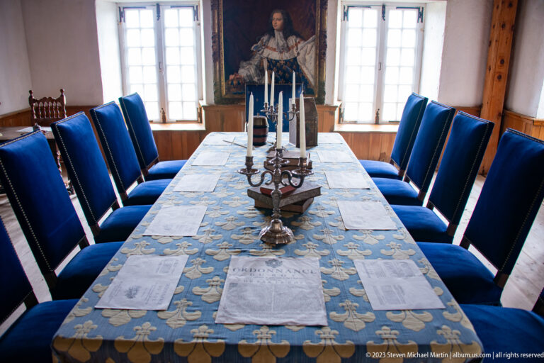 Long blue-covered dining table lined with high-backed blue chairs, candlesticks, napkins, tall windows, and a portrait.