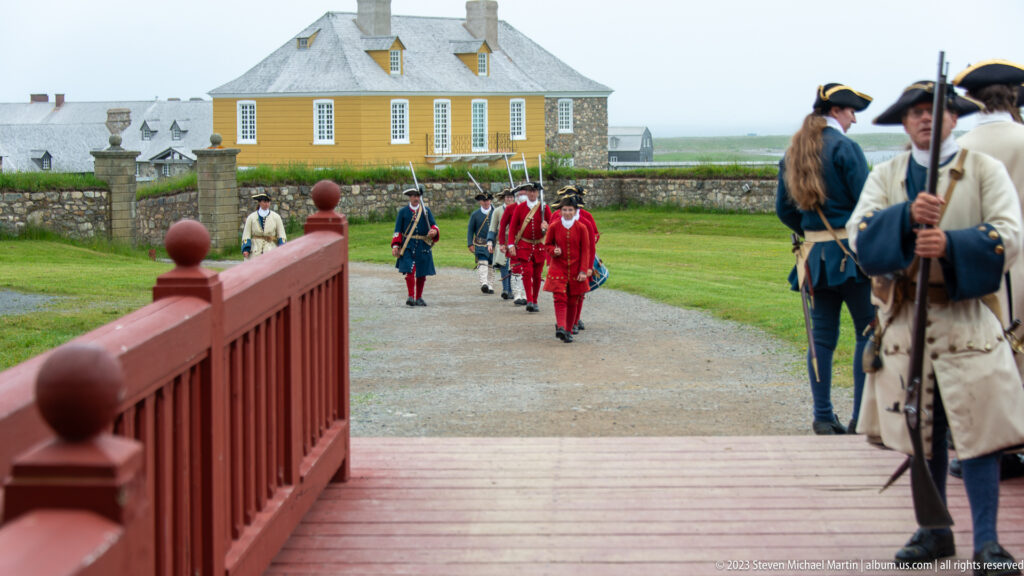 Costumed reenactors in red military coats and pale period dress walk along a gravel path beside reconstructed buildings.