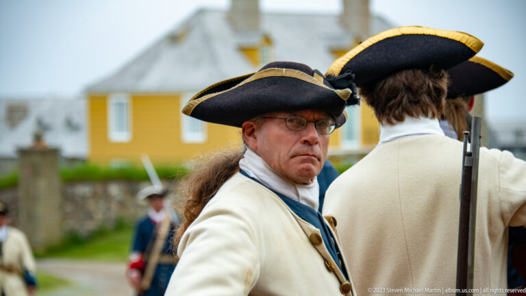 Close view of a reenactor in tricorn hat, pale coat, and spectacles beside another interpreter carrying a long firearm.