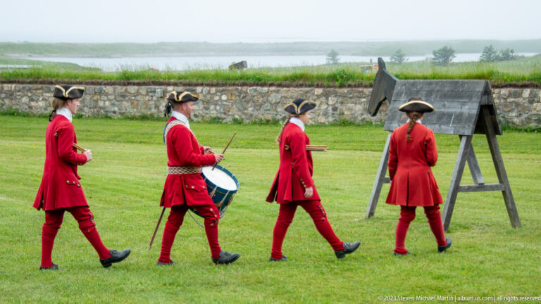 Four red-coated reenactors walk across grass, including a drummer, with a stone wall and misty water in the background.