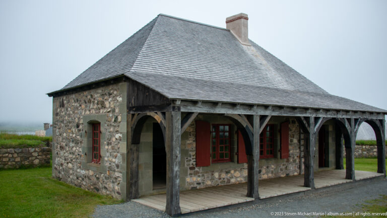 Small reconstructed stone building with steep hipped roof, timber veranda, red shutters, multipane windows, and chimney.