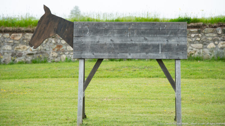 Weathered wooden horse-shaped structure with plank body and braced legs standing on grass before a low stone wall.