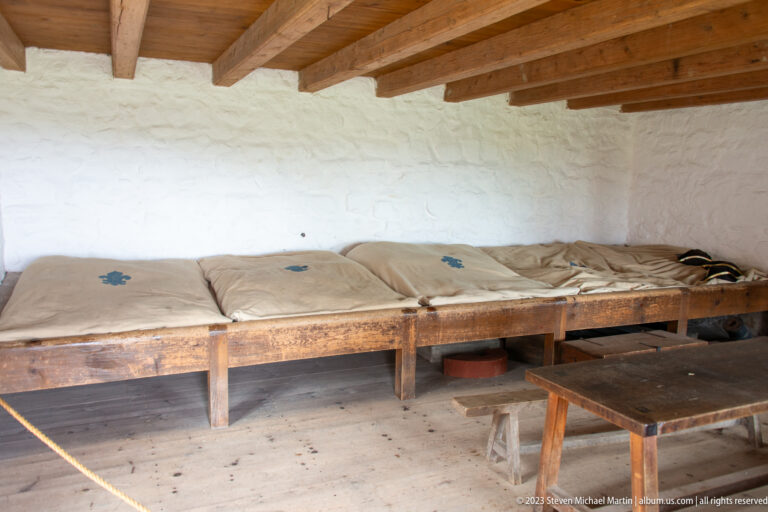 Long wooden sleeping platforms with thin bedding line a whitewashed wall beneath exposed beams in reconstructed communal quarters.