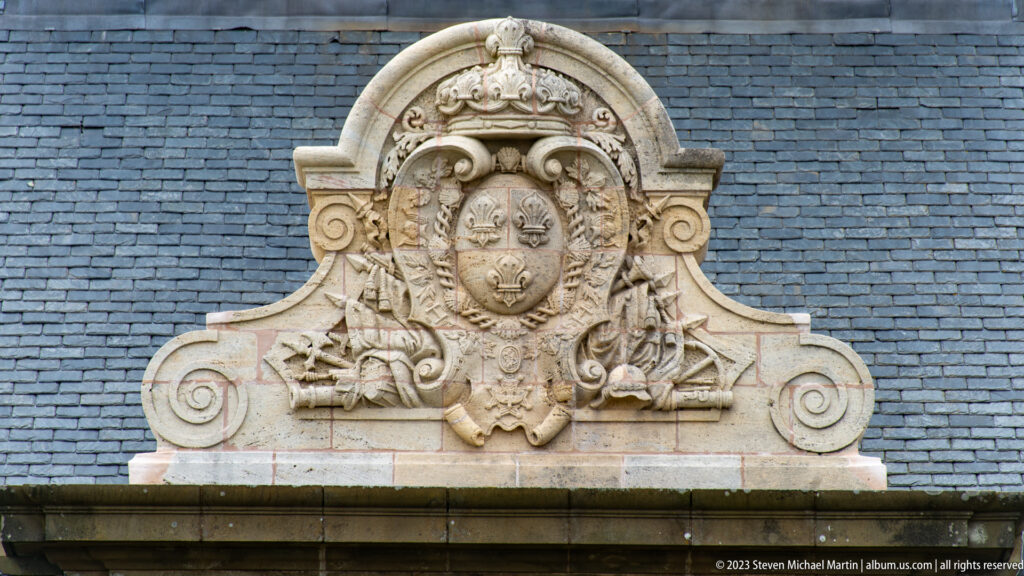 Carved stone royal arms with crown, fleur-de-lis shield, scrollwork, and military trophies set against a slate roof.