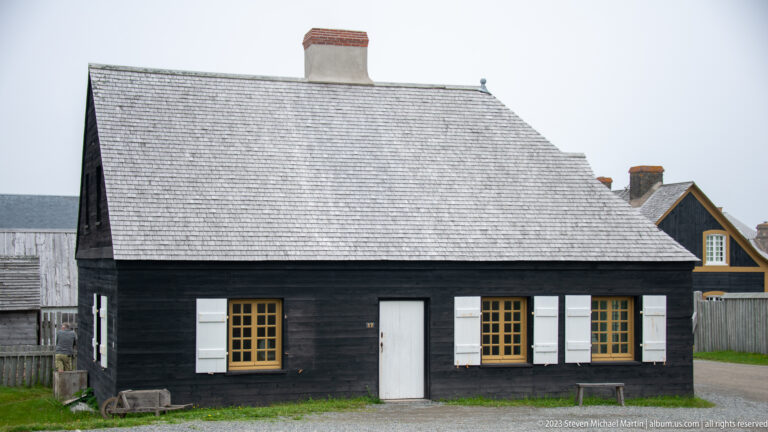 Black-painted timber house with central white door, yellow-trimmed windows, white shutters, and steep shingled roof.