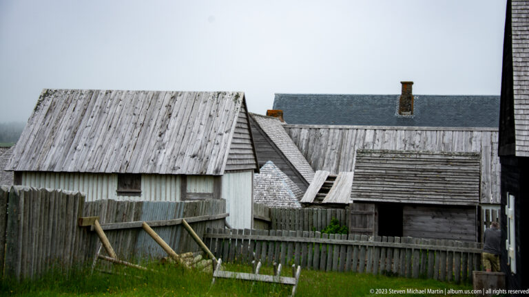 Cluster of weathered wooden sheds and fences with steep roofs in a reconstructed service yard at Louisbourg.
