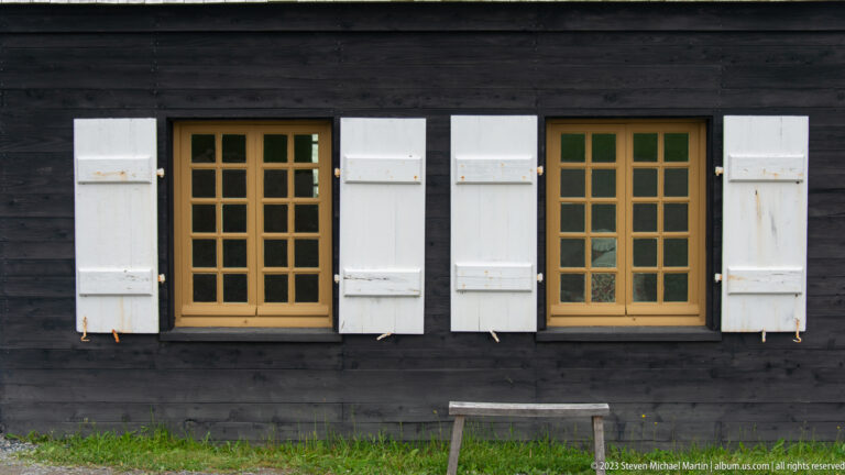 Two ochre-trimmed multipane windows with white shutters set into a black-painted timber wall.