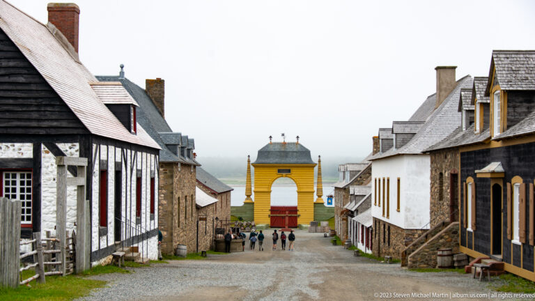 Gravel street lined with reconstructed timber and stone houses leading toward a bright yellow gate and distant visitors.