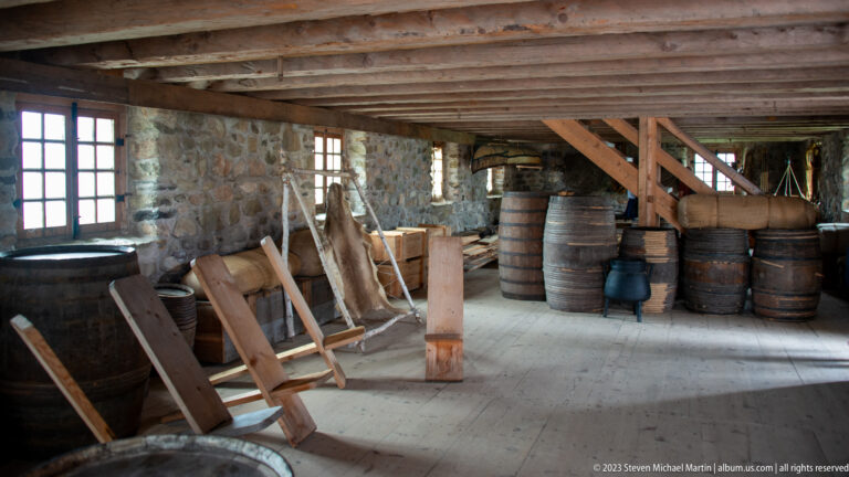 Rough stone-and-timber interior filled with wooden barrels, tubs, planks, and work equipment in a reconstructed storage space.
