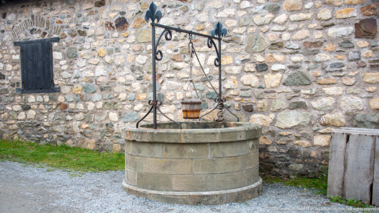 Circular stone well with wrought-iron pulley frame and hanging bucket beside a rough stone wall.