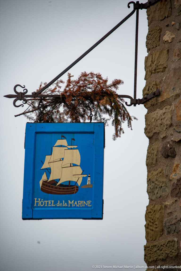 Blue Hôtel de la Marine hanging sign with painted sailing ship, suspended from an ornate iron bracket on a stone wall.