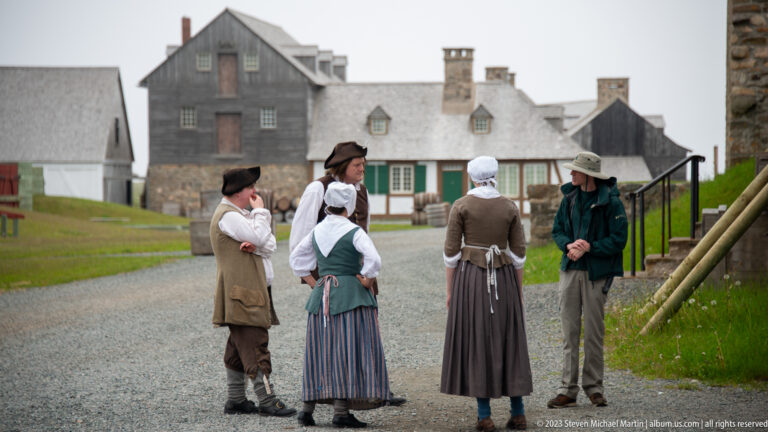 Four costumed interpreters in eighteenth-century civilian clothing talk with a modern visitor on a gravel street among reconstructed buildings.