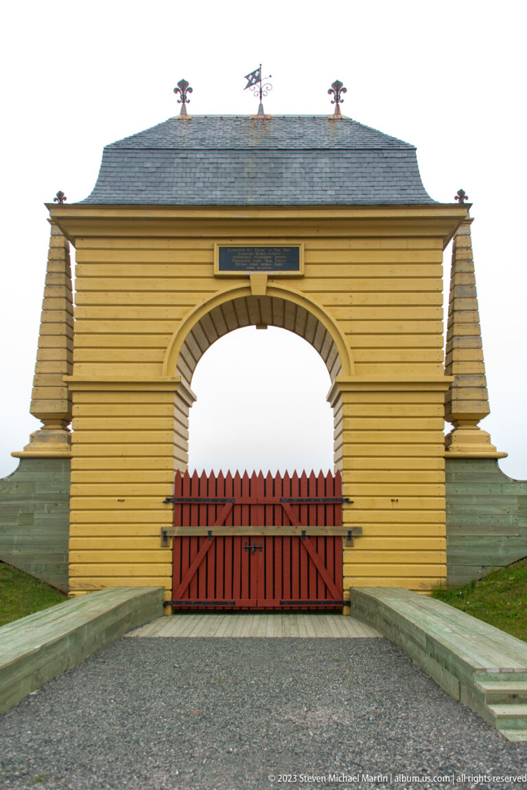 Bright yellow timber gateway with central arch, closed red double doors, sloping side walls, and decorative roofline ornaments.