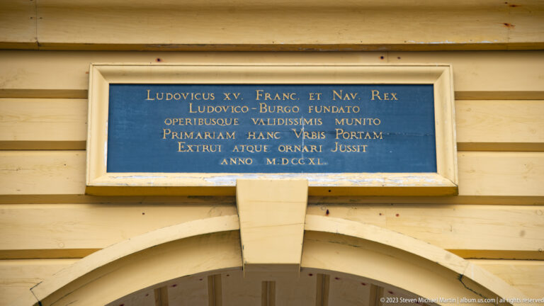 Blue plaque with gold Latin inscription naming Louis XV mounted above the arch of a yellow timber gateway.