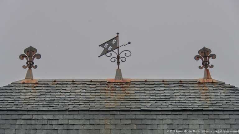 Three dark metal roof finials, including fleur-de-lis-shaped ornaments, mounted above a gray slate roof.