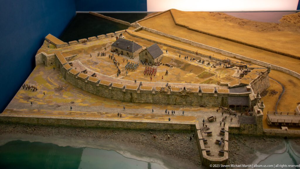 Overhead view of a detailed scale model with walls, buildings, troops, and waterfront.