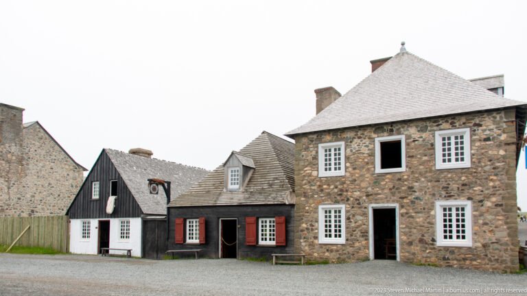 Group of reconstructed stone and timber houses with white windows, red shutters, steep roofs, and narrow spacing.