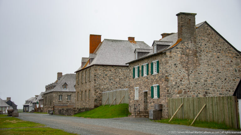 Stone buildings with steep roofs, chimneys, green shutters, and timber fences line a gravel street inside Louisbourg.