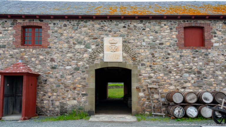 Stone warehouse façade with a central passage, red doors, carved sign, and stacked wooden barrels.