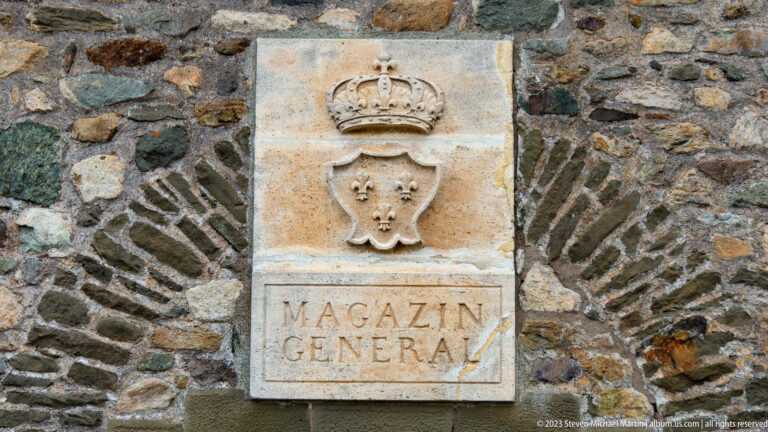 Carved stone MAGAZIN GENERAL plaque with crown and fleur-de-lis shield set into rough masonry.