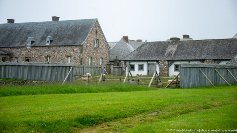 Grassy fenced service yard with stone buildings, shingled roofs, gates, and stacked lumber.