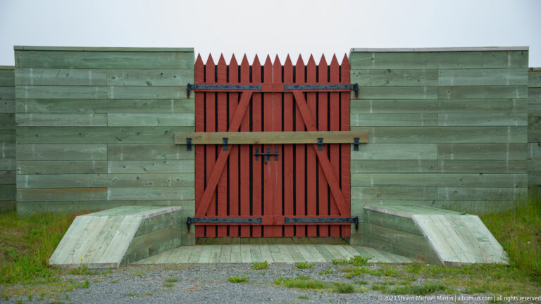 Red braced wooden gate with pointed pickets set into a green plank defensive barrier.