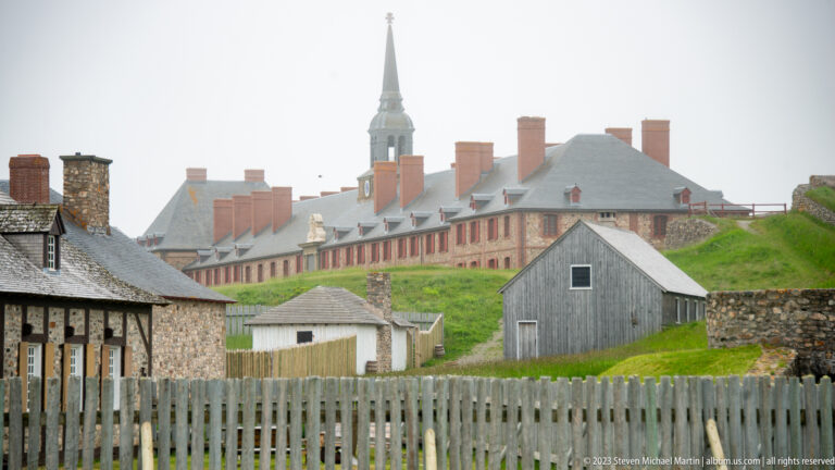 Long red-trimmed barracks and chapel spire rise beyond fences and outbuildings at Louisbourg.