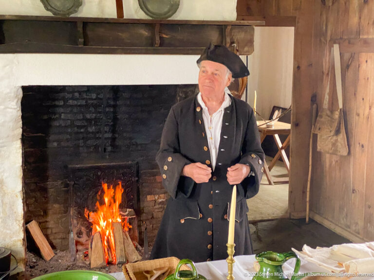 Costumed male interpreter stands beside a burning open hearth in a reconstructed historic interior.