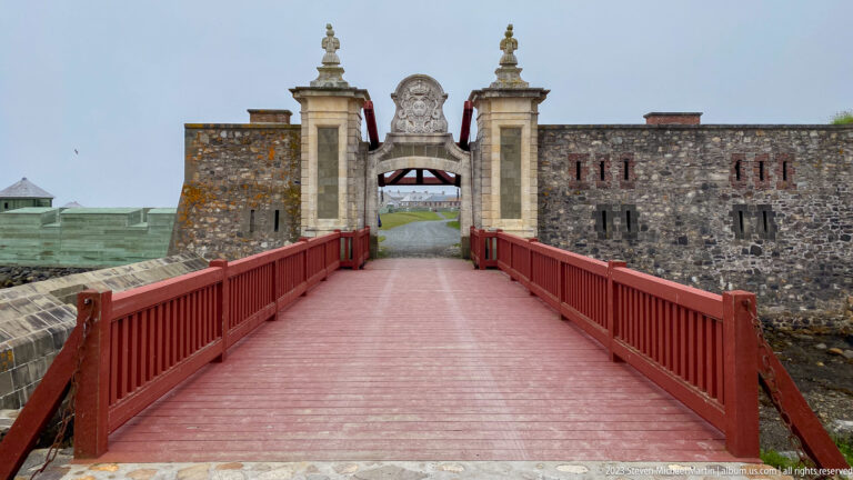 Red wooden bridge aligned with an ornate stone gateway between fortress walls at Louisbourg.