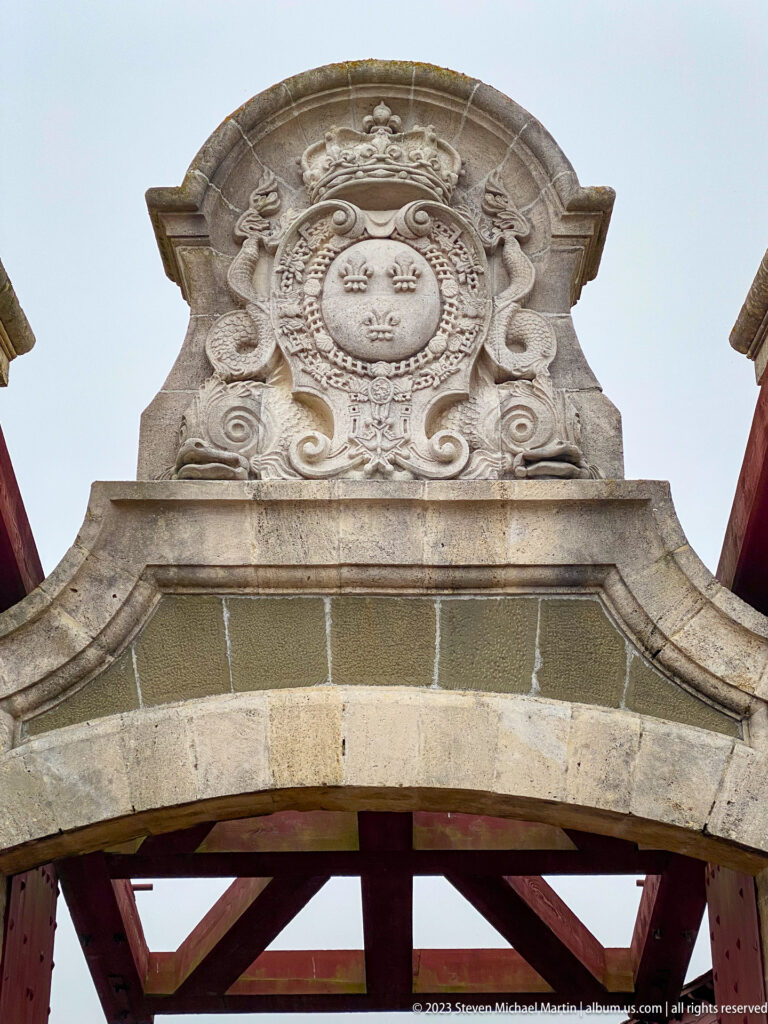 Carved crowned shield with fleur-de-lis and scrollwork above a stone archway.