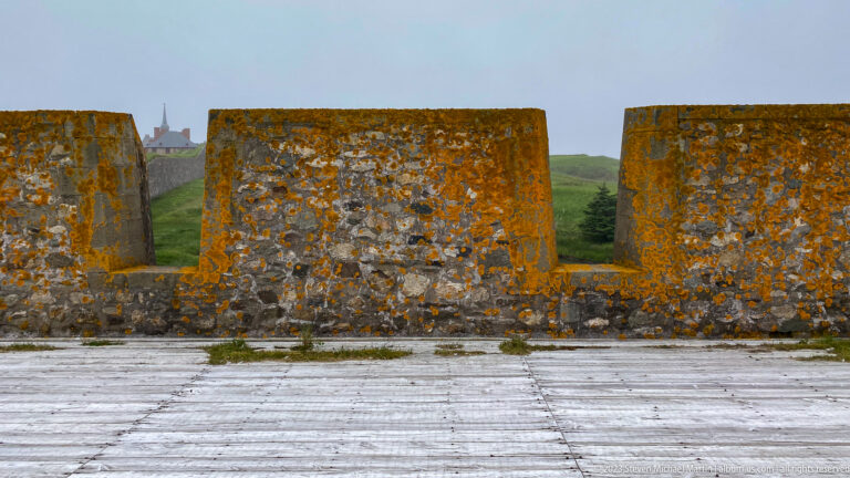Lichen-covered stone parapet with rectangular embrasures and distant fortress buildings visible through one opening.
